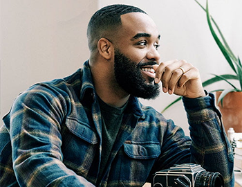 A bearded man sits at a table, focused on a camera in front of him.