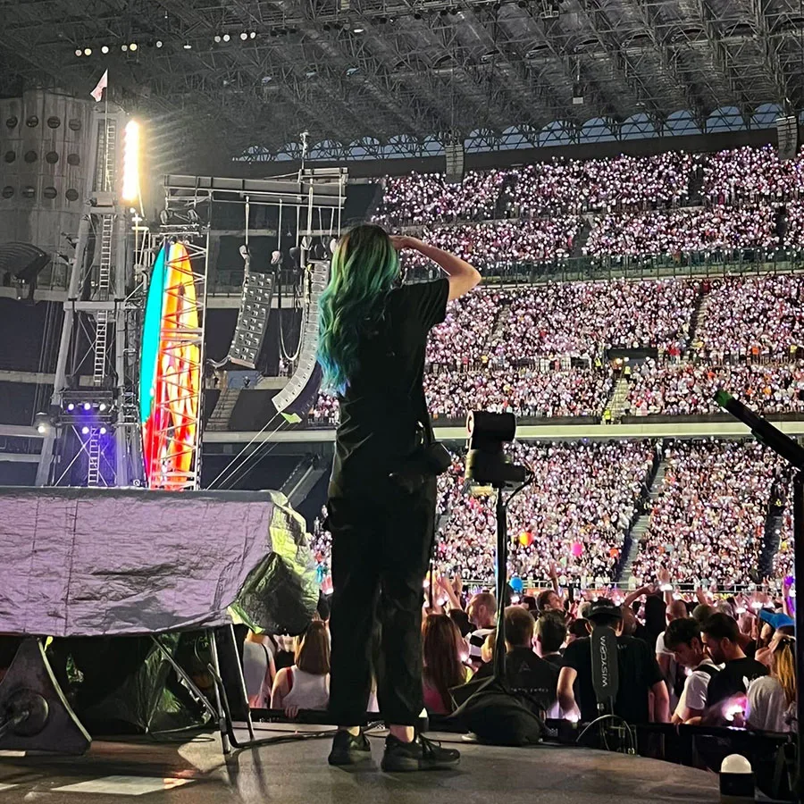 A person with blue hair stands on stage, addressing a large crowd of enthusiastic audience members.