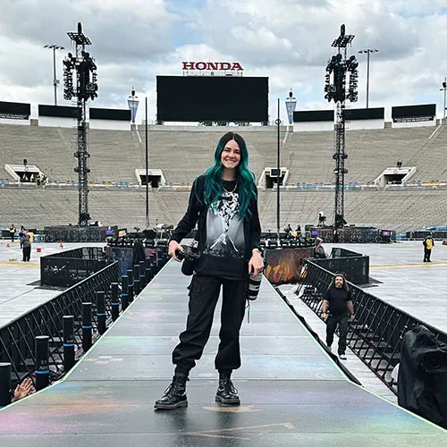 A woman with green hair stands confidently on a stage, engaging the audience with her presence.