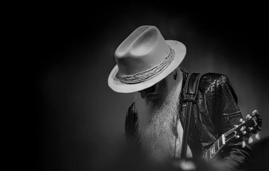 A man with a long beard and hat playing an acoustic guitar, focused on his music in a casual setting.
