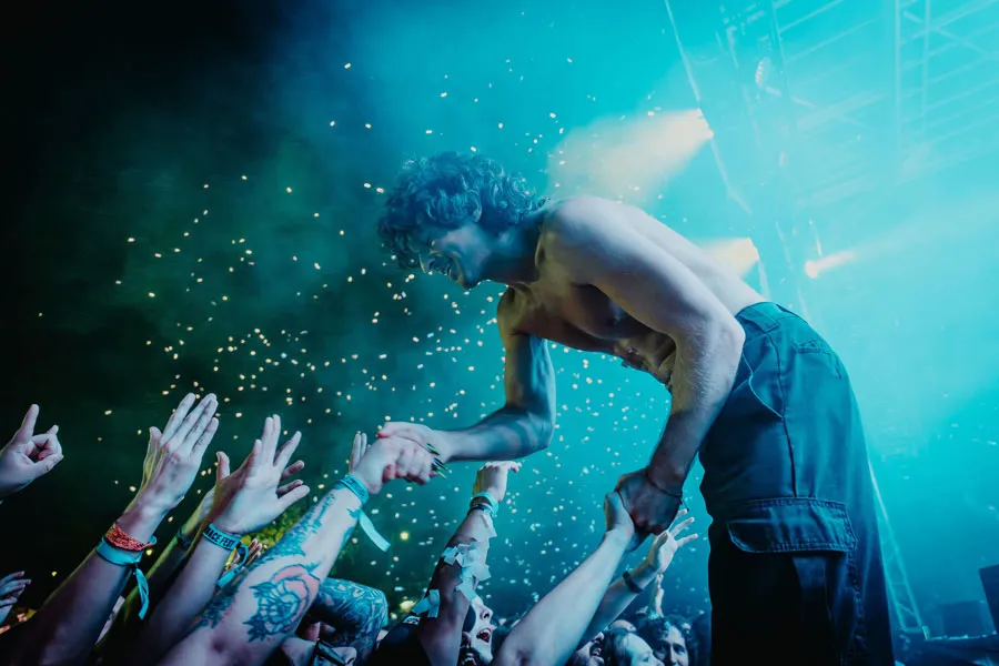 A shirtless man stands on stage with his hands raised, engaging the audience with enthusiasm.
