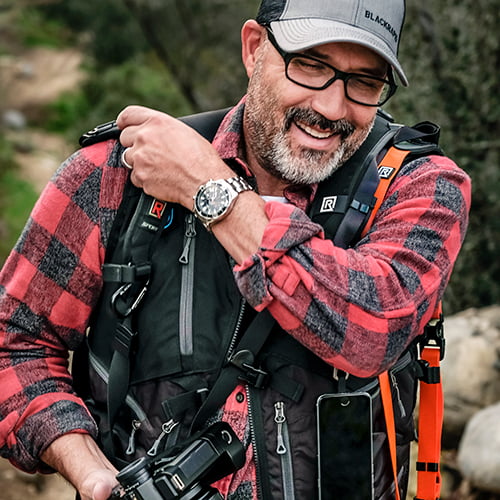 A smiling man with a backpack and camera, ready for an adventure outdoors.