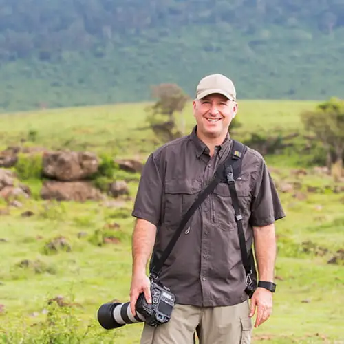 A man holding a camera stands in a grassy field, ready to capture the surrounding landscape.