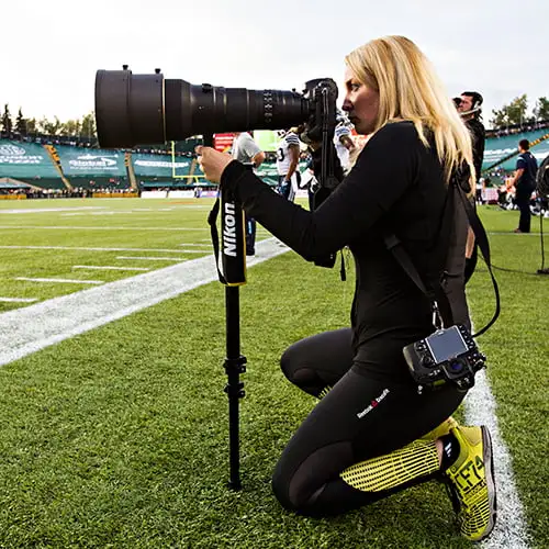A woman kneels on a grassy field, holding a camera and preparing to take a photograph.