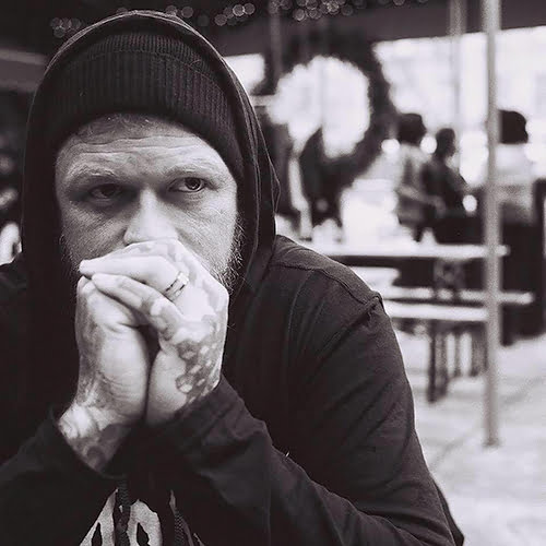 A tattooed man sits at a table, resting his chin on his hands, deep in thought.