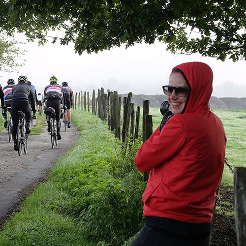 A woman in a red jacket stands beside a group of cyclists, smiling and engaging with them outdoors.