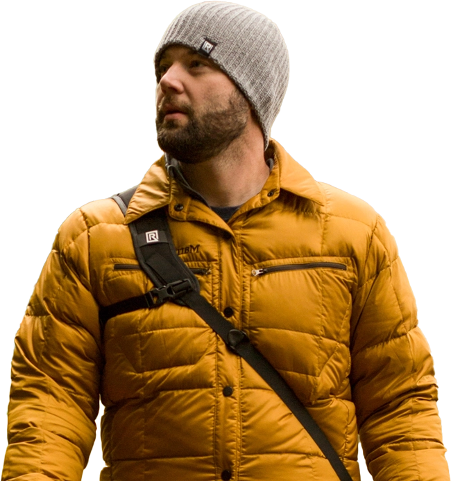 A man wearing a gray knit beanie and a mustard yellow puffer jacket stands in a forest