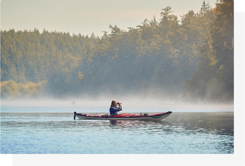 Person sitting in a red kayak on a calm lake, using binoculars while surrounded by mist and dense evergreen forest in the background using Blackrapid best camera strap.